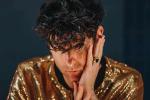 A person with curly dark hair, wearing a shiny gold sequin jacket, rests their face in one hand and looks directly at the camera against a dark background.
