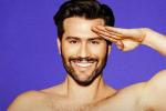 A smiling man with dark hair and a beard salutes with his right hand against a bright purple background. He is shirtless and looking directly at the camera.