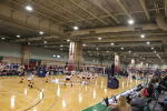A wide view of an indoor sports facility with multiple volleyball courts. Several teams are playing matches while spectators watch from the bleachers along the sidelines. The ceiling is high with visible beams and lighting.