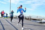 A woman in athletic gear runs on a boardwalk, smiling and making peace signs with both hands. Other runners are visible behind her. The sky is clear and blue.