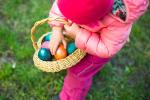 A young child wearing a pink jacket and hat holds a woven basket filled with colorful Easter eggs while standing on grass.