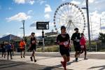 Runners participating in a race on a paved path, with a large Ferris wheel and partly cloudy sky in the background. Some runners wear race bibs and athletic clothing.