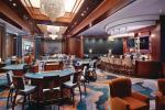 Spacious casino interior with gaming tables, slot machines, a long bar with stools, modern lighting, and a large chandelier. The decor features warm wood tones, soft seating, and a patterned carpet.