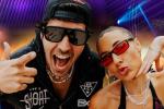 Two people wearing dark sunglasses and trendy outfits pose energetically at a party with colorful lights in the background; one is making a peace sign, and both are smiling and playful.
