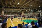 A basketball game is in progress on an indoor court, with players actively moving and spectators seated around the court watching the action under bright overhead lights.