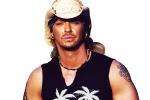 A man with long blond hair wears a light cowboy hat, sleeveless black shirt with palm tree graphics, a necklace, and has a tattoo on his upper arm. He is posing against a white background.
