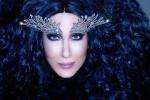 A person with long, voluminous black hair wears dramatic, silver, wing-shaped eye adornments and has bold makeup, looking directly at the camera against a dark background.