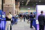 People walk and interact at an indoor convention or trade show, with booths and displays lining both sides of a wide aisle under a high ceiling.