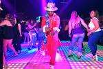 A man in a bright red suit and hat dances on a colorful, illuminated floor, surrounded by people dancing in a lively nightclub with purple lighting.