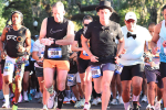 A group of runners at the starting line of a race, some wearing costumes such as a black top hat and bow tie, and brightly colored athletic gear, with trees and sunlight in the background.