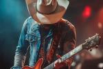 A musician wearing a cowboy hat and denim jacket plays an electric guitar on stage, with colorful lights and a blurred background.
