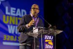 A person speaks passionately at a podium with a microphone, wearing glasses and a lanyard. Behind them, a large screen displays the text “LEAD UNITE WIN 32BJ SEIU” in bold letters.