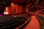 A spacious, modern theater with rows of empty seats, red carpeting, and curved balconies, all illuminated by soft overhead lighting. The stage is set with a blank screen and purple lighting.