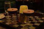 Two espresso martini cocktails with frothy tops sit on a checkered table alongside a glass of water and a candle in a yellow patterned holder.