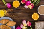 Breakfast scene with croissants on a plate, orange juice, coffee, orange halves, and a loaf of bread on a wooden table, with pink and yellow tulips arranged around the food.